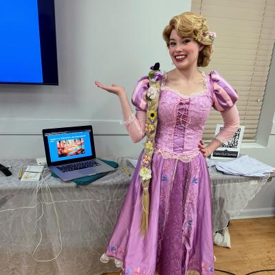 Brianna Williamson hosts Disney Trivia Night in our Actor's Hall. Brianna poses next to her trivia slidedeck, dressed as Princess Rapunzel, with a long blonde braid and a bright purple dress. 