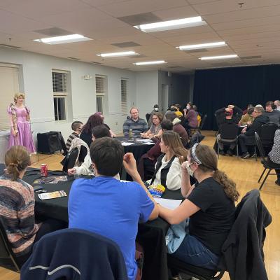 Brianna Williamson, dressed as Rapunzel, hosts Disney Trivia Night in our Actor's Hall. Players of all ages sit around tables with black tablecloths, preparing their answer to the next trivia question.