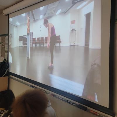 Nikki Mirza demonstrates a slow roll-up movement over Zoom. She is projected on a large projector screen. 