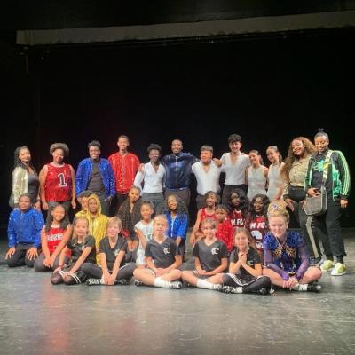 Young dancers from After School Dance Fund's Tentacion, Centerstage Academy for the Arts' Showbiz Next Generation, and the Culkin School of Irish Dance stand grouped together, smiling at the camera. They wear vibrant costumes of all colors in front of the darkened stage.
