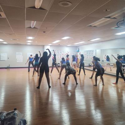 In the background, students dance, spread out in a rehearsal room. They are half-lit and half-silhouetted.