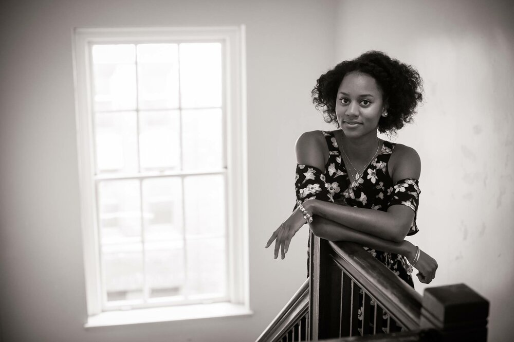 A grayscale photo of Simone Brown leaning on the banister of a stairwell. She wears a floral dress, bracelets, and has short curly natural hair. Light streams in from a window behind her.